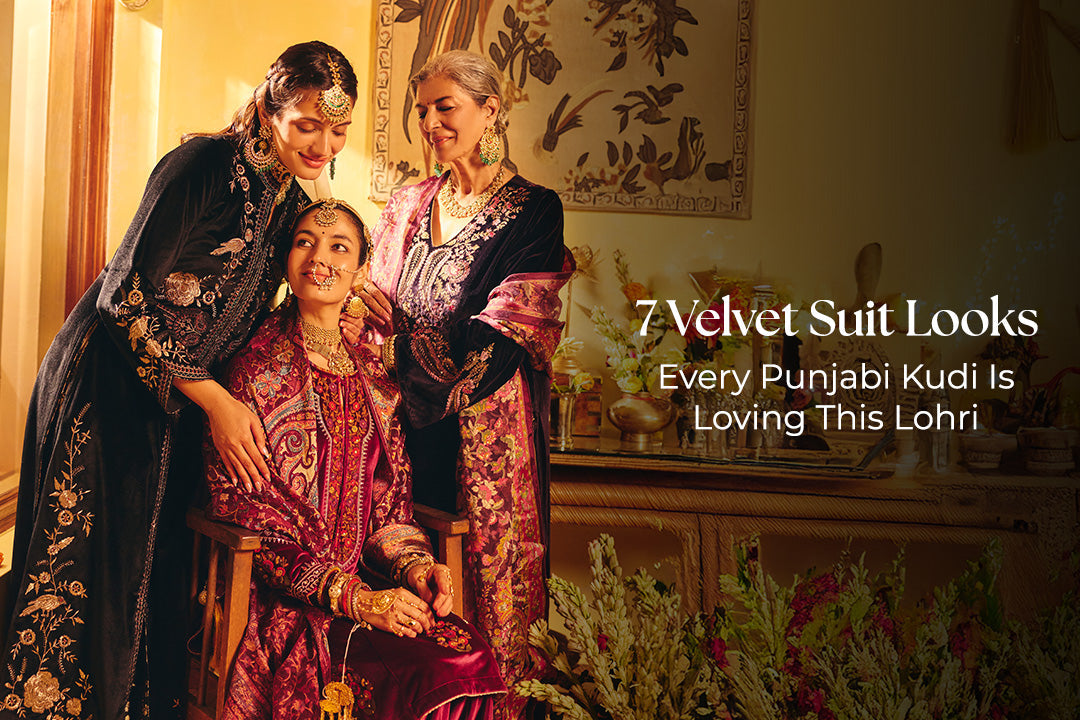 Three Punjabi women dressed in embroidered velvet suit sets in rich maroon and jewel tones, celebrating Lohri together in a festive indoor setting